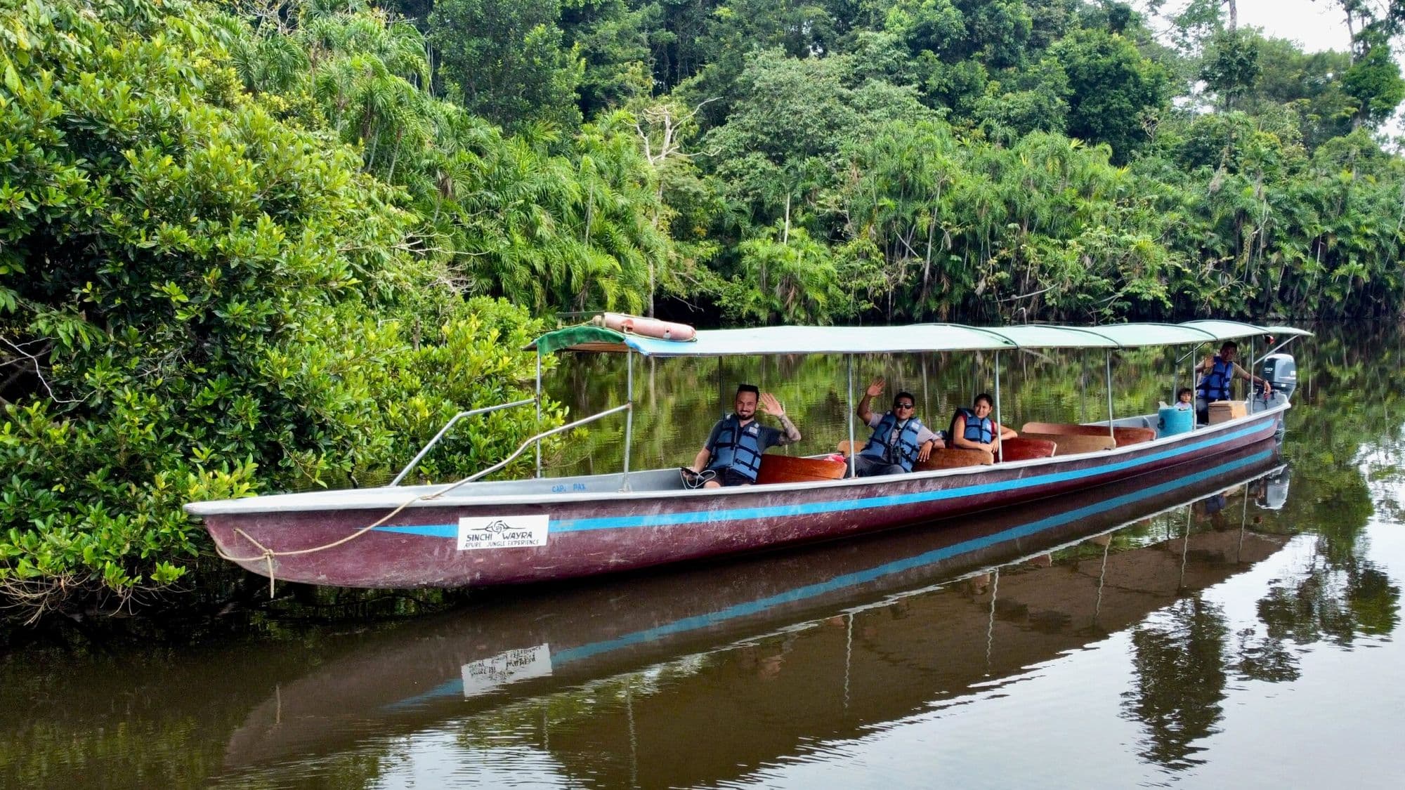 Turistas saludando desde un bote con toldo de Sinchi Wayra en un río tranquilo del Amazonas, Ecuador.
