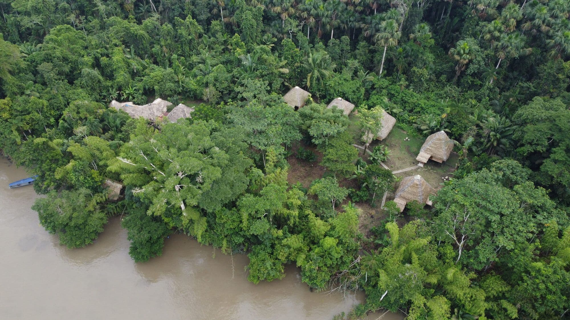 Aerial drone view of Sinchi Wayra Lodge: thatched-roof cabins in a clearing of the Amazon rainforest beside the brown river